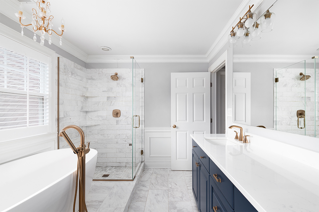 Bathroom with grey walls, white crown molding, glass shower, bath tub, and navy cabinets with gold fixtures